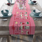 The pink Christmas table runner features the AriesCat Christmas Cocktails design. It features different midcentury style cocktails artwork with a red square present bow around them. 