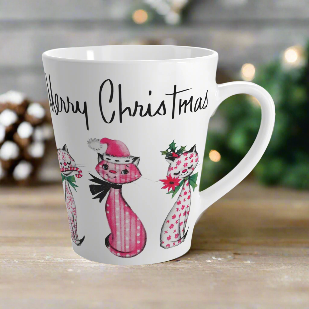 This ceramic latte coffee mug says Merry Christmas in midcentury lettering and features 3 adorable midcentury retro Christmas cats. They are pink with different patterns. They each have a bow around their neck, one is holding a candy cane in their mouth, one is wearing a pink santa hat and one has a flower in her mouth. Nostalgic and adorable. 