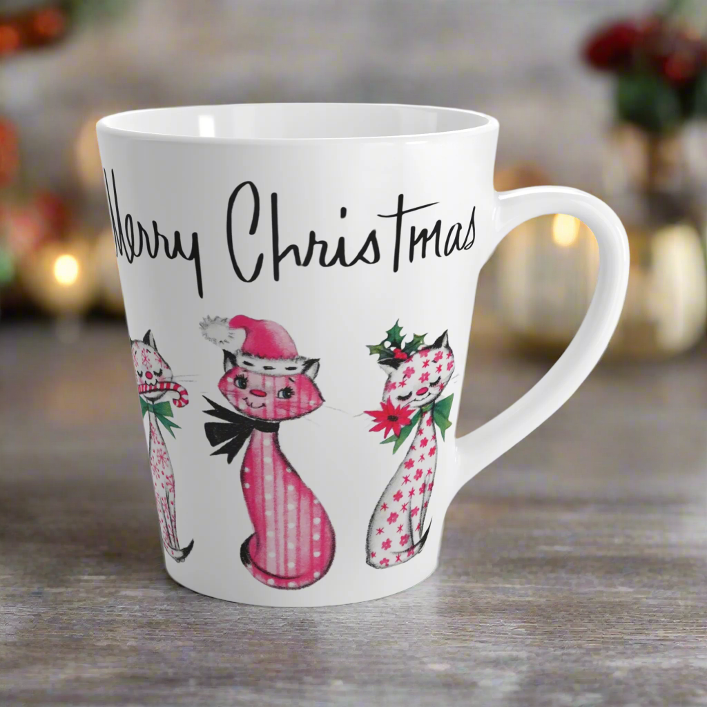 This ceramic latte coffee mug says Merry Christmas in midcentury lettering and features 3 adorable midcentury retro Christmas cats. They are pink with different patterns. They each have a bow around their neck, one is holding a candy cane in their mouth, one is wearing a pink santa hat and one has a flower in her mouth. Nostalgic and adorable. 