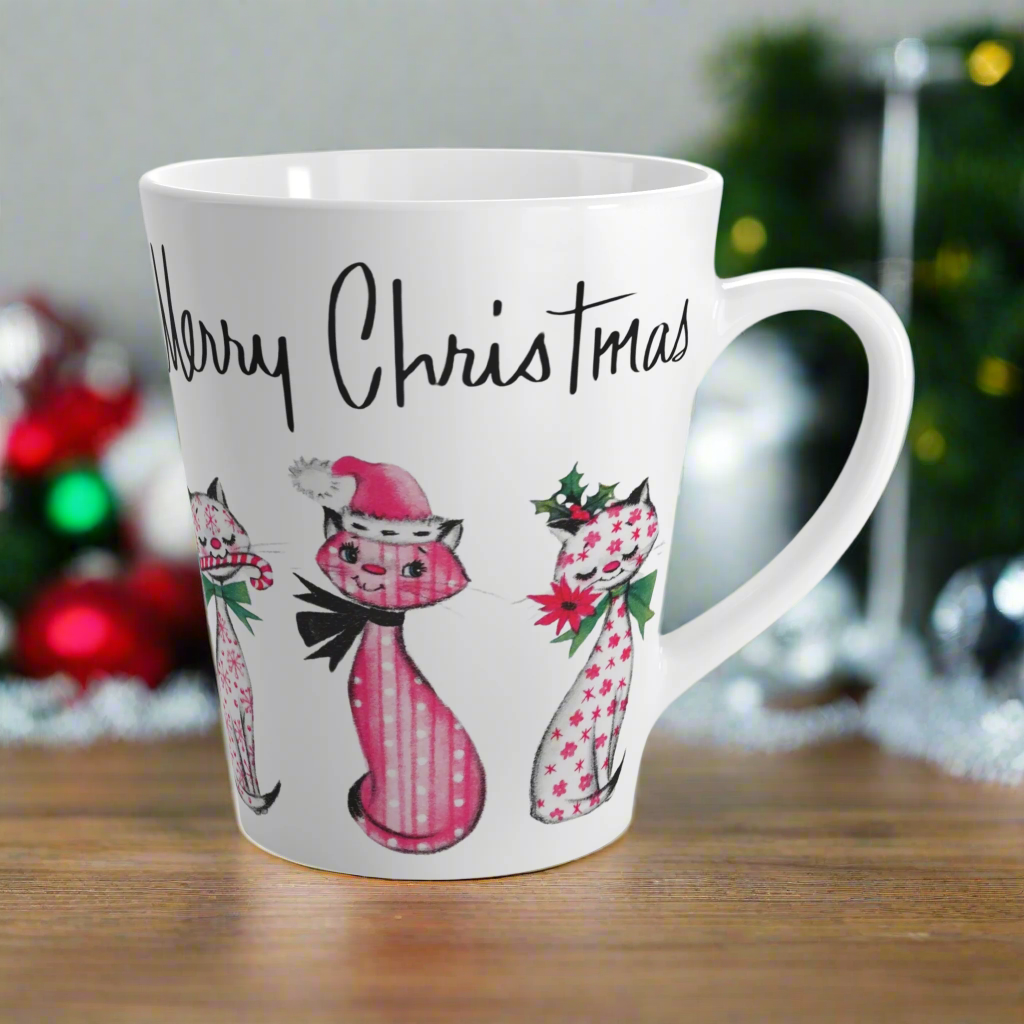 This ceramic latte coffee mug says Merry Christmas in midcentury lettering and features 3 adorable midcentury retro Christmas cats. They are pink with different patterns. They each have a bow around their neck, one is holding a candy cane in their mouth, one is wearing a pink santa hat and one has a flower in her mouth. Nostalgic and adorable. 