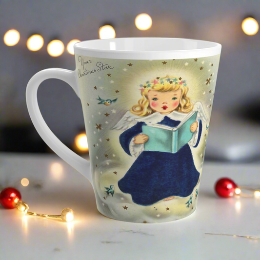 This glossy ceramic Christmas latte mug features a vintage illustration of a blond angel holding a blue book and is singing. There are bluebirds and stars surrounding her. She is wearing a star crown.