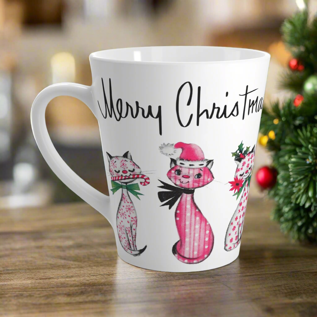 This ceramic latte coffee mug says Merry Christmas in midcentury lettering and features 3 adorable midcentury retro Christmas cats. They are pink with different patterns. They each have a bow around their neck, one is holding a candy cane in their mouth, one is wearing a pink santa hat and one has a flower in her mouth. Nostalgic and adorable. 
