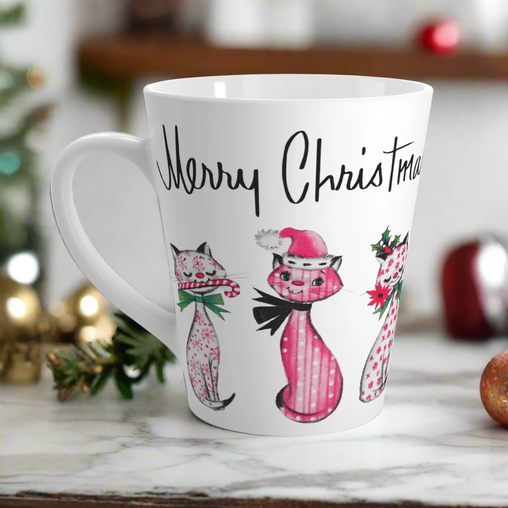 This ceramic latte coffee mug says Merry Christmas in midcentury lettering and features 3 adorable midcentury retro Christmas cats. They are pink with different patterns. They each have a bow around their neck, one is holding a candy cane in their mouth, one is wearing a pink santa hat and one has a flower in her mouth. Nostalgic and adorable. 