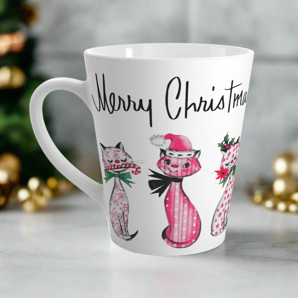 This ceramic latte coffee mug says Merry Christmas in midcentury lettering and features 3 adorable midcentury retro Christmas cats. They are pink with different patterns. They each have a bow around their neck, one is holding a candy cane in their mouth, one is wearing a pink santa hat and one has a flower in her mouth. Nostalgic and adorable. 