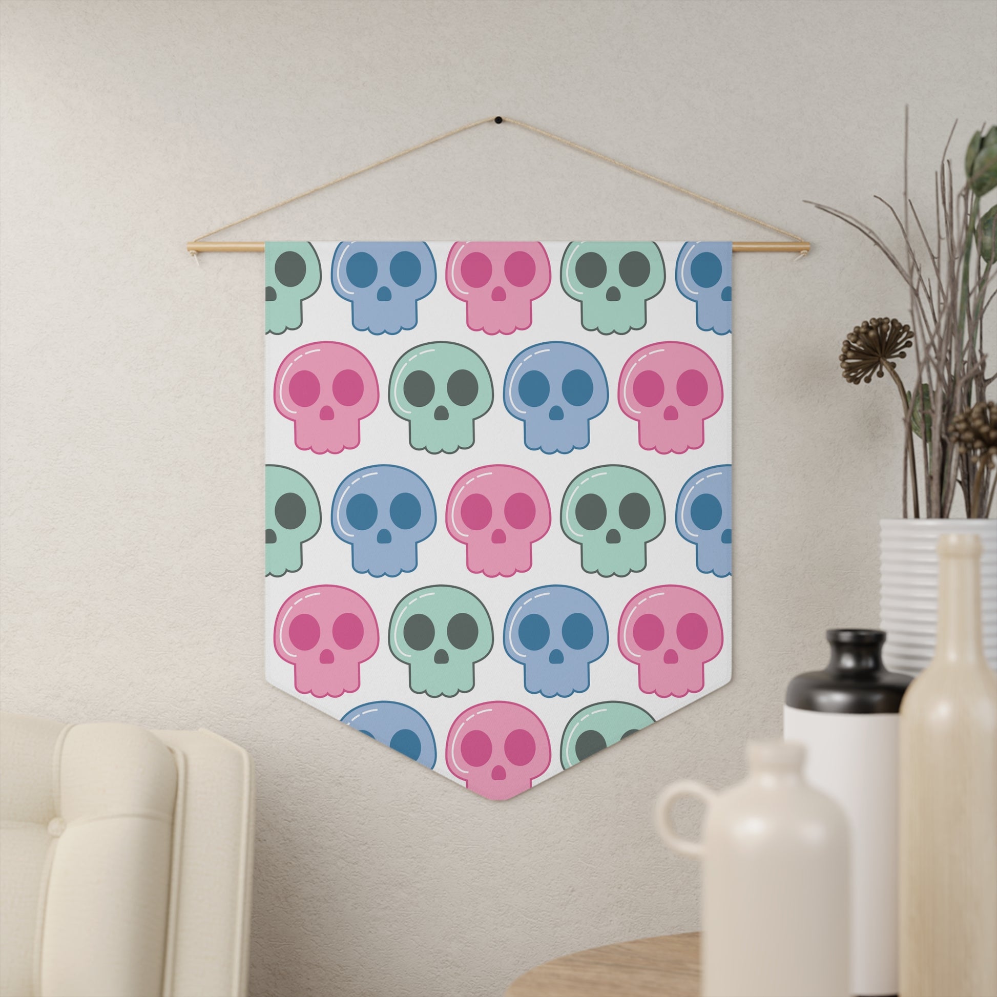 This pennant banner wall hanging features cute pastel goth skulls patterned in pink, green and blue with a white background on fabric canvas. It hands on a wooden dowel.