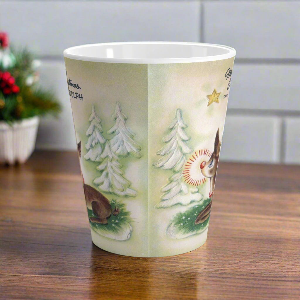 This ceramic latte coffee mug features a midcentury vintage Christmas illustration featuring Rudolph the Red Nosed Reindeer sitting down looking up at a gold star in the sky surrounded by snowy trees. It says: Merry Christmas Says Rudolph.