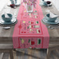 The pink Christmas table runner features the AriesCat Christmas Cocktails design. It features different midcentury style cocktails artwork with a red square present bow around them. 