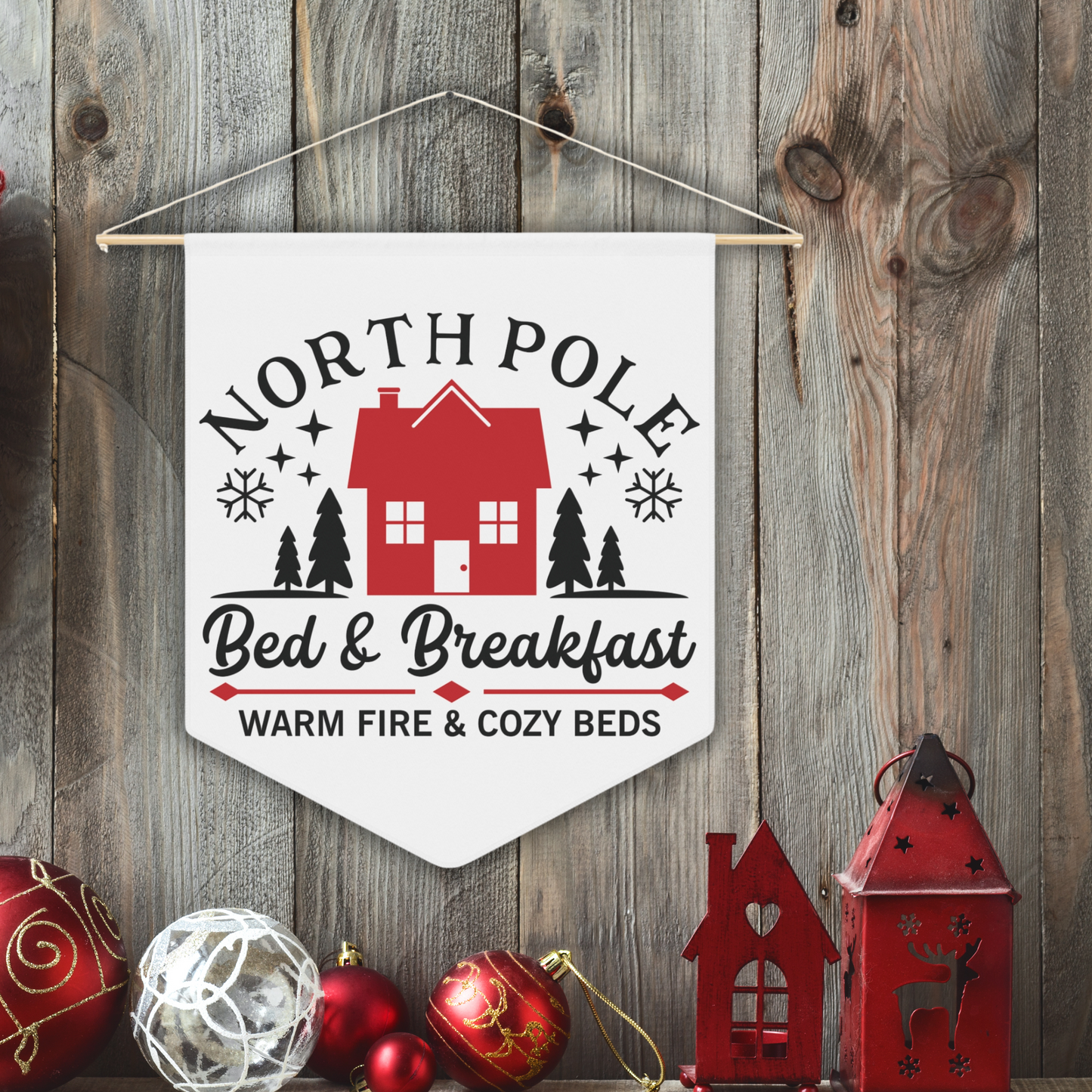 This white Christmas wall hanging pennant banner flag says North Pole Bed and Breakfast Warm Fire and Cozy Beds. It features a red house with trees and stars.