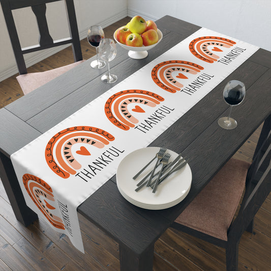 This white table runner features an orange boho rainbow with black hearts and doodles, there is an orange heart in the center. Beneath the rainbow it says Thankful in black lettering.