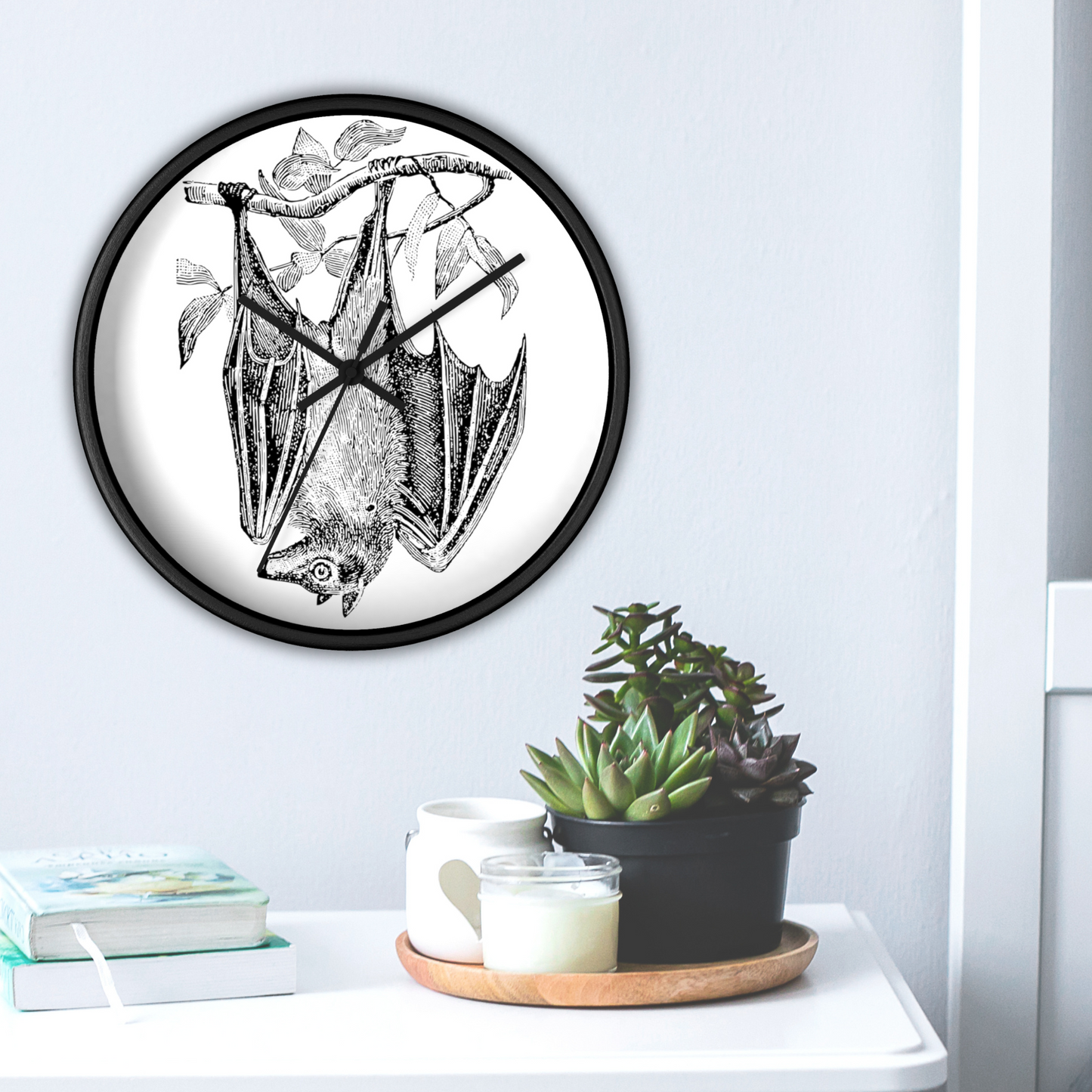 This black wall clock features a vintage illustration of a bat hanging upside down from a branch. The frame and hands of the clock are black.