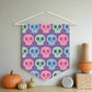 This fabric wall hanging features pastel skulls in pink, green and blue with a purple background. They are staggered in a pattern.