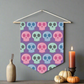 This fabric wall hanging features pastel skulls in pink, green and blue with a purple background. They are staggered in a pattern.