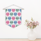 This pennant banner wall hanging features cute pastel goth skulls patterned in pink, green and blue with a white background on fabric canvas. It hands on a wooden dowel.