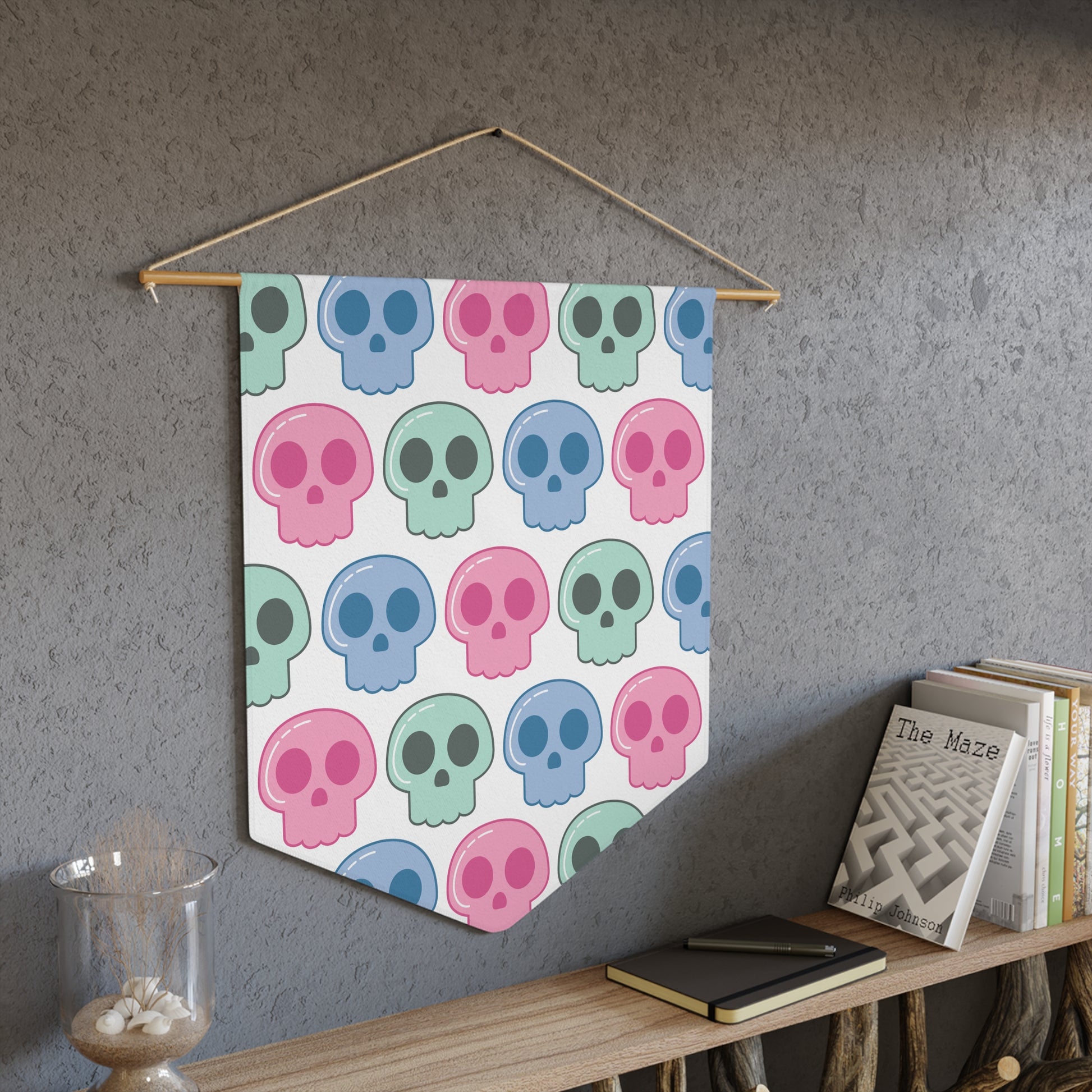 This pennant banner wall hanging features cute pastel goth skulls patterned in pink, green and blue with a white background on fabric canvas. It hands on a wooden dowel.