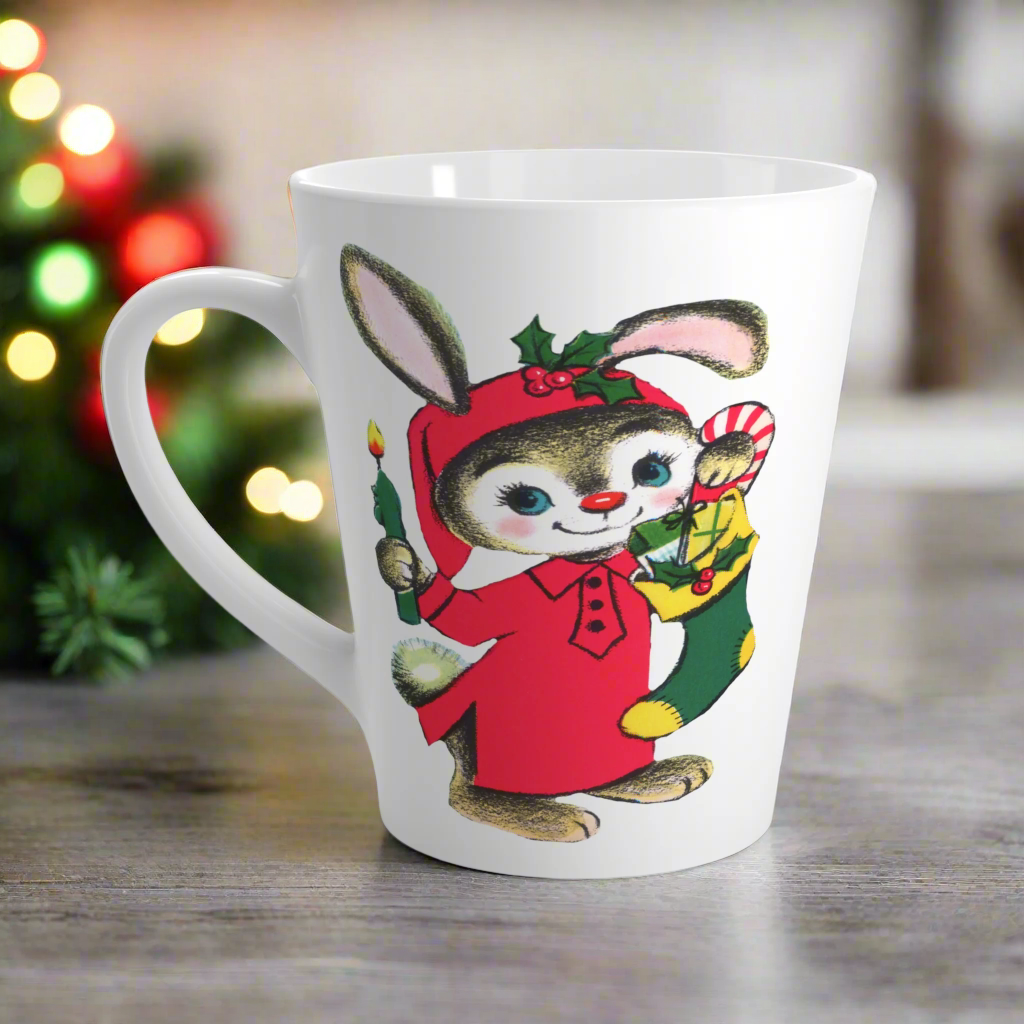 This ceramic glossy latte coffee mug features a midcentury Christmas illustration of an adorable bunny. The bunny is wearing a red sleeping cap and nightgown. He is holding a candle in one hand and a green and yellow christmas stocking with presents.