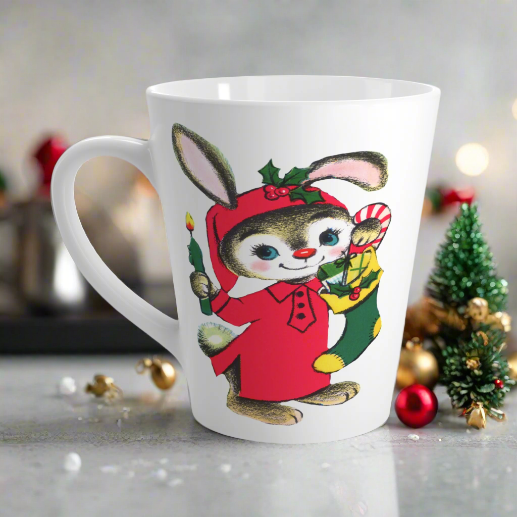 This ceramic glossy latte coffee mug features a midcentury Christmas illustration of an adorable bunny. The bunny is wearing a red sleeping cap and nightgown. He is holding a candle in one hand and a green and yellow christmas stocking with presents.