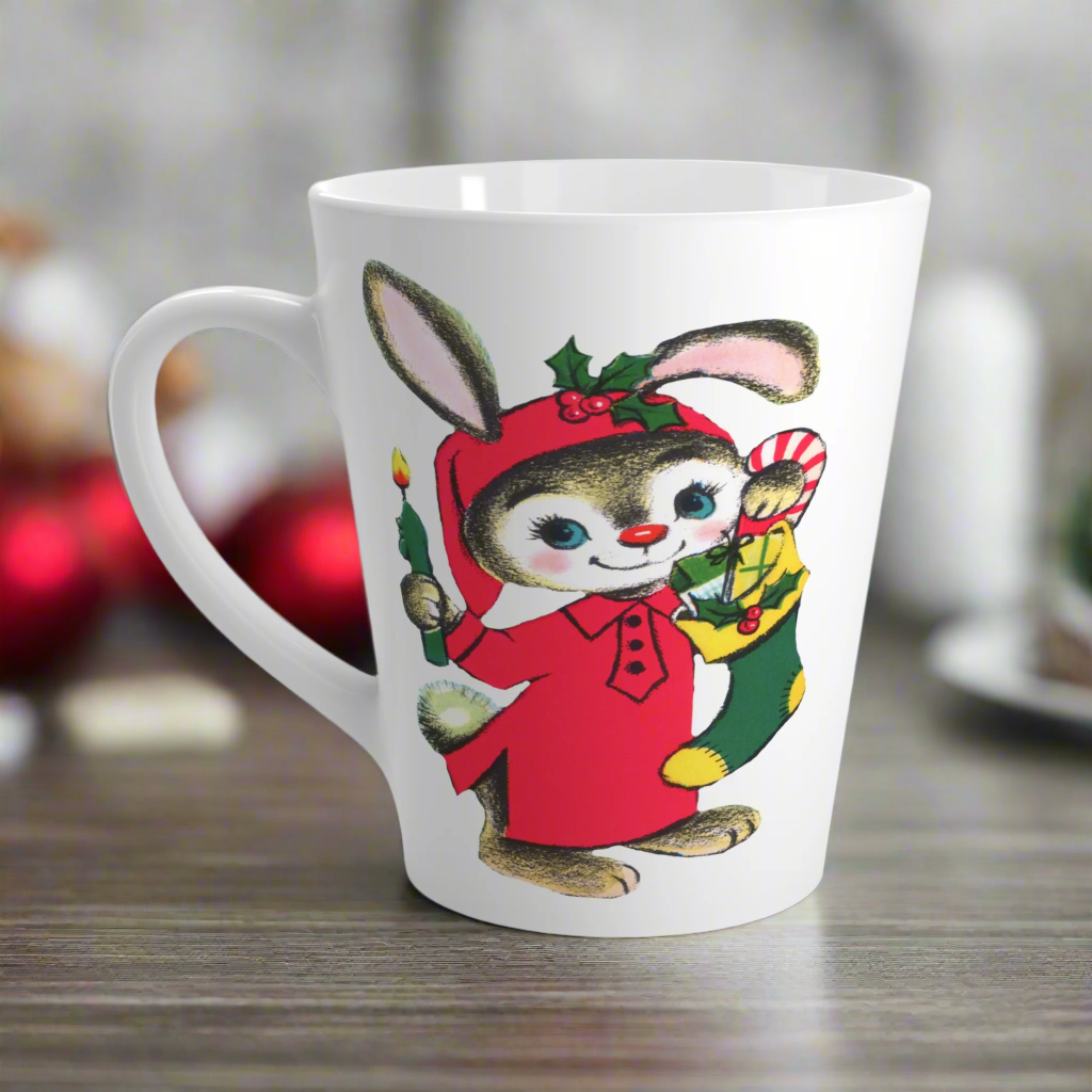 This ceramic glossy latte coffee mug features a midcentury Christmas illustration of an adorable bunny. The bunny is wearing a red sleeping cap and nightgown. He is holding a candle in one hand and a green and yellow christmas stocking with presents.