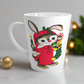 This ceramic glossy latte coffee mug features a midcentury Christmas illustration of an adorable bunny. The bunny is wearing a red sleeping cap and nightgown. He is holding a candle in one hand and a green and yellow christmas stocking with presents.