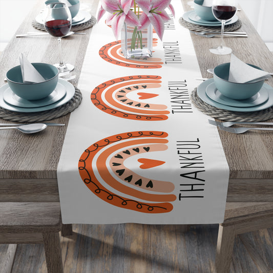 This white table runner features an orange boho rainbow with black hearts and doodles, there is an orange heart in the center. Beneath the rainbow it says Thankful in black lettering.