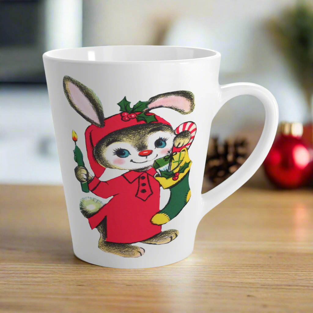 This ceramic glossy latte coffee mug features a midcentury Christmas illustration of an adorable bunny. The bunny is wearing a red sleeping cap and nightgown. He is holding a candle in one hand and a green and yellow christmas stocking with presents.