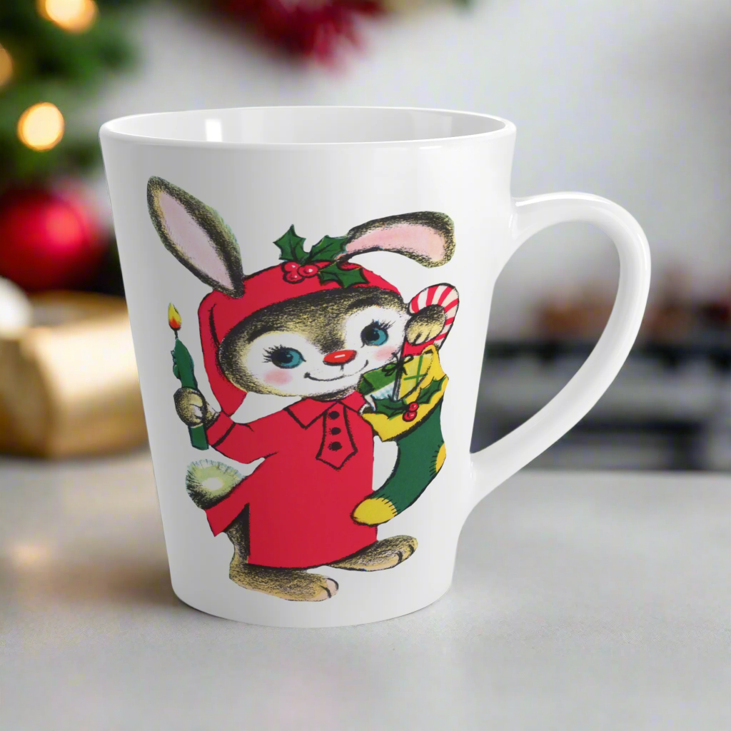 This ceramic glossy latte coffee mug features a midcentury Christmas illustration of an adorable bunny. The bunny is wearing a red sleeping cap and nightgown. He is holding a candle in one hand and a green and yellow christmas stocking with presents.