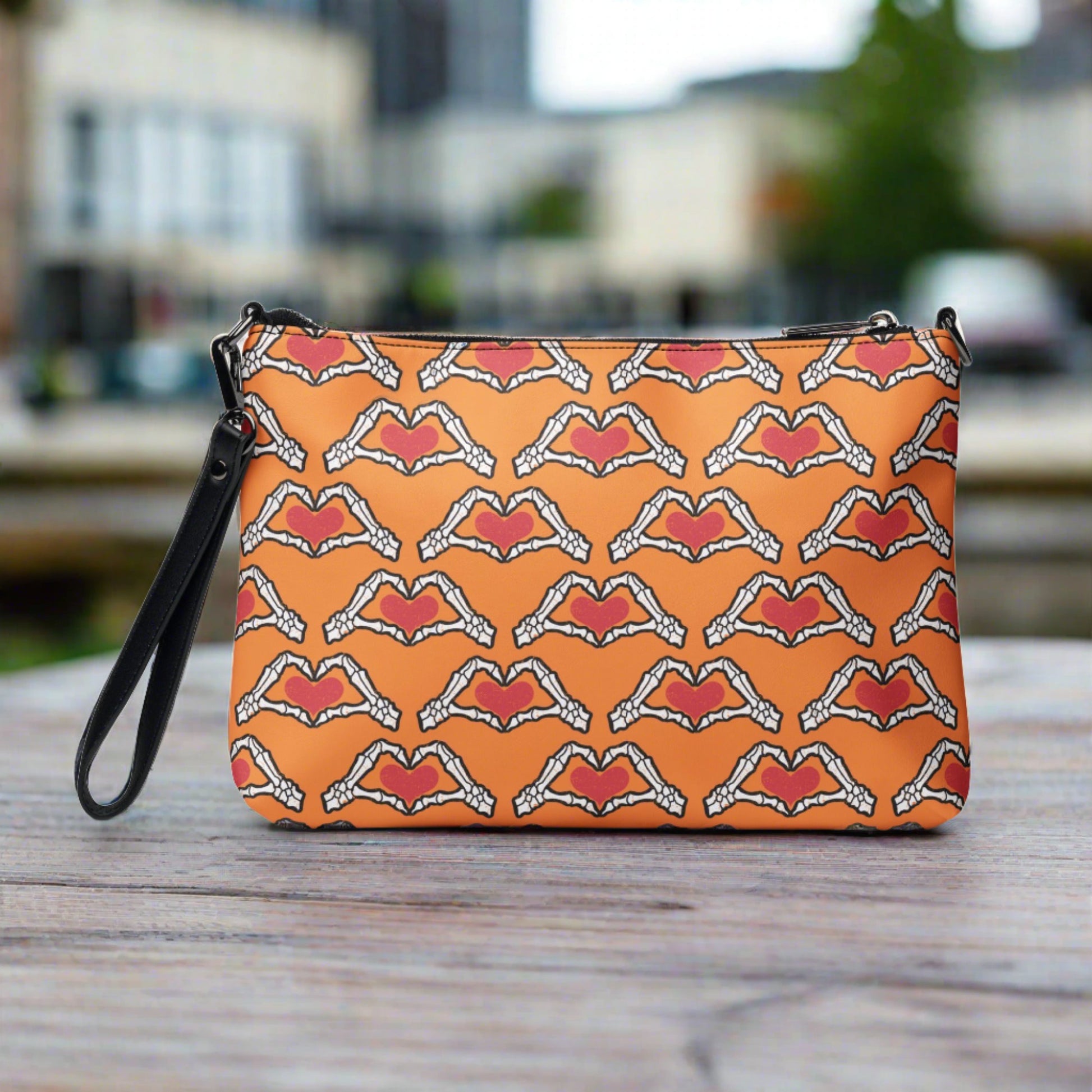 This orange crossbody purse converts to a large wristlet clutch. It features a distressed print of skeleton hands forming a heart with a red heart inside. One side the design is large and the other is a smaller patterned print.