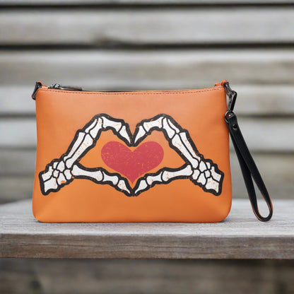 This orange crossbody purse converts to a large wristlet clutch. It features a distressed print of skeleton hands forming a heart with a red heart inside. One side the design is large and the other is a smaller patterned print.