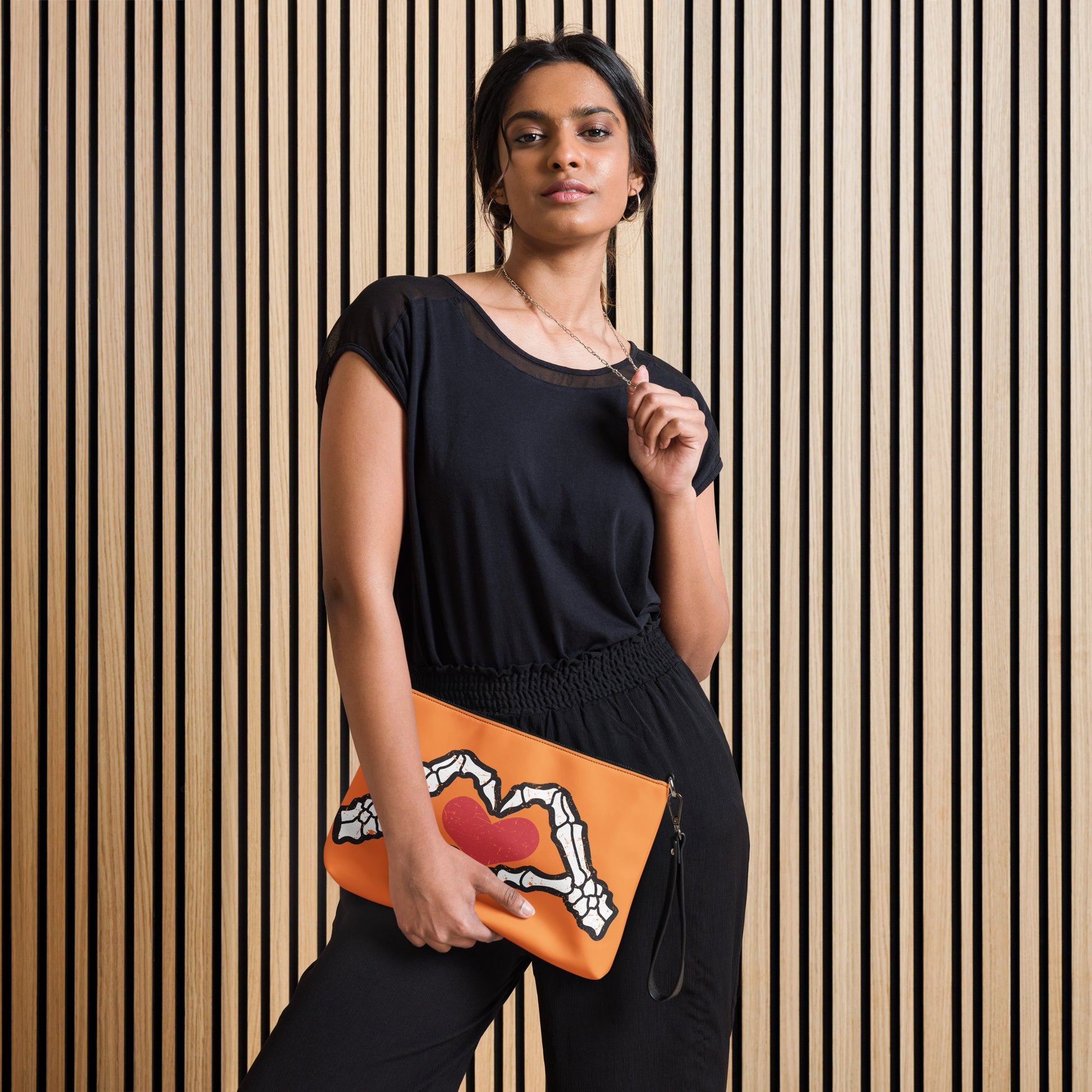 This orange crossbody purse converts to a large wristlet clutch. It features a distressed print of skeleton hands forming a heart with a red heart inside. One side the design is large and the other is a smaller patterned print.