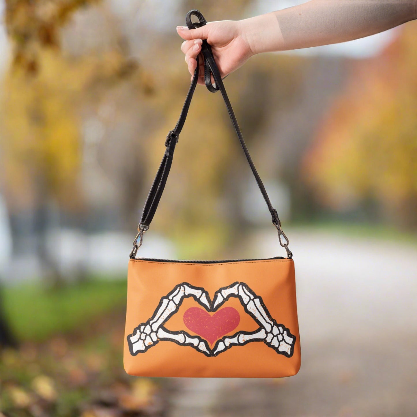 This orange crossbody purse converts to a large wristlet clutch. It features a distressed print of skeleton hands forming a heart with a red heart inside. One side the design is large and the other is a smaller patterned print.