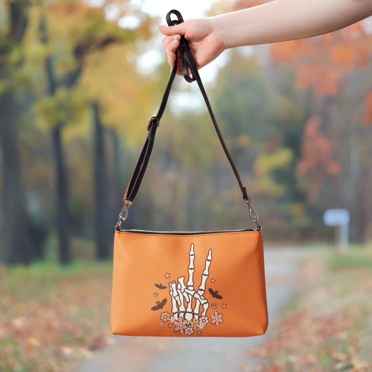 This orange crossbody purse features a skeleton hand making a peace sign surrounded by bats, flowers and peace signs.