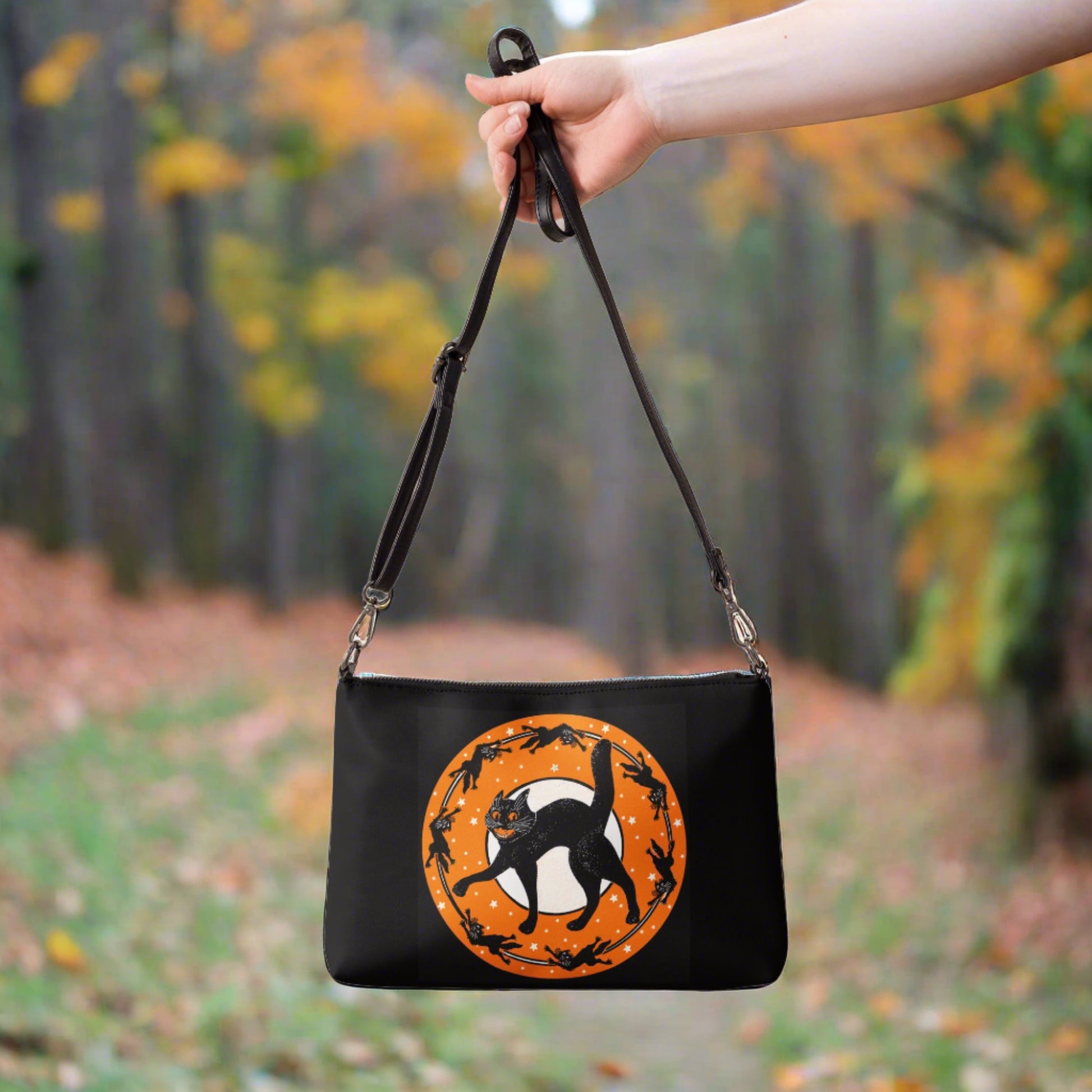 This black crossbody pursefeatures a retro Halloween print of a black cat with little black cats surrounding him in a circle with an orange circle with white stars around the moon.