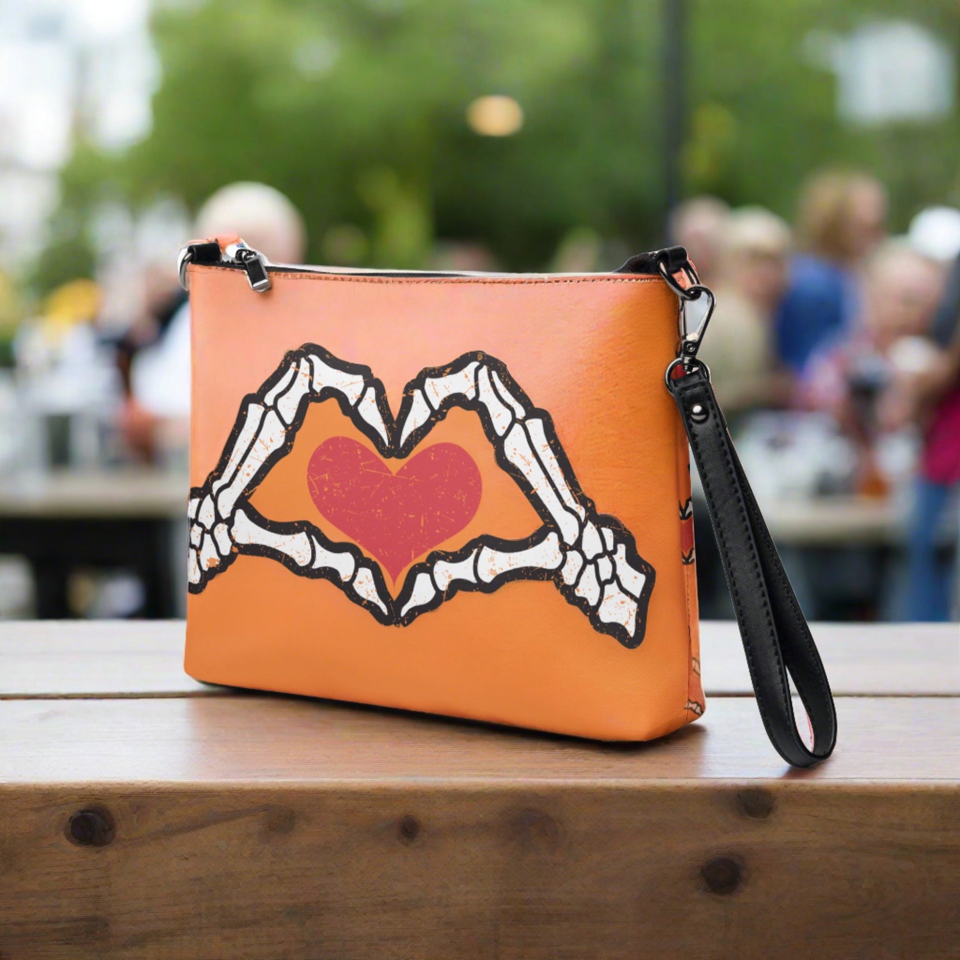 This orange crossbody purse converts to a large wristlet clutch. It features a distressed print of skeleton hands forming a heart with a red heart inside. One side the design is large and the other is a smaller patterned print.