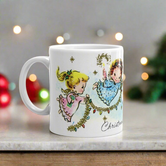 This ceramic glossy coffee mug features a midcentury Christmas illustration that says Christmas Joy and features three young angels flying and decorating a Christmas tree with tinsel and lights. There is a blond, brunette, and redhead angel.