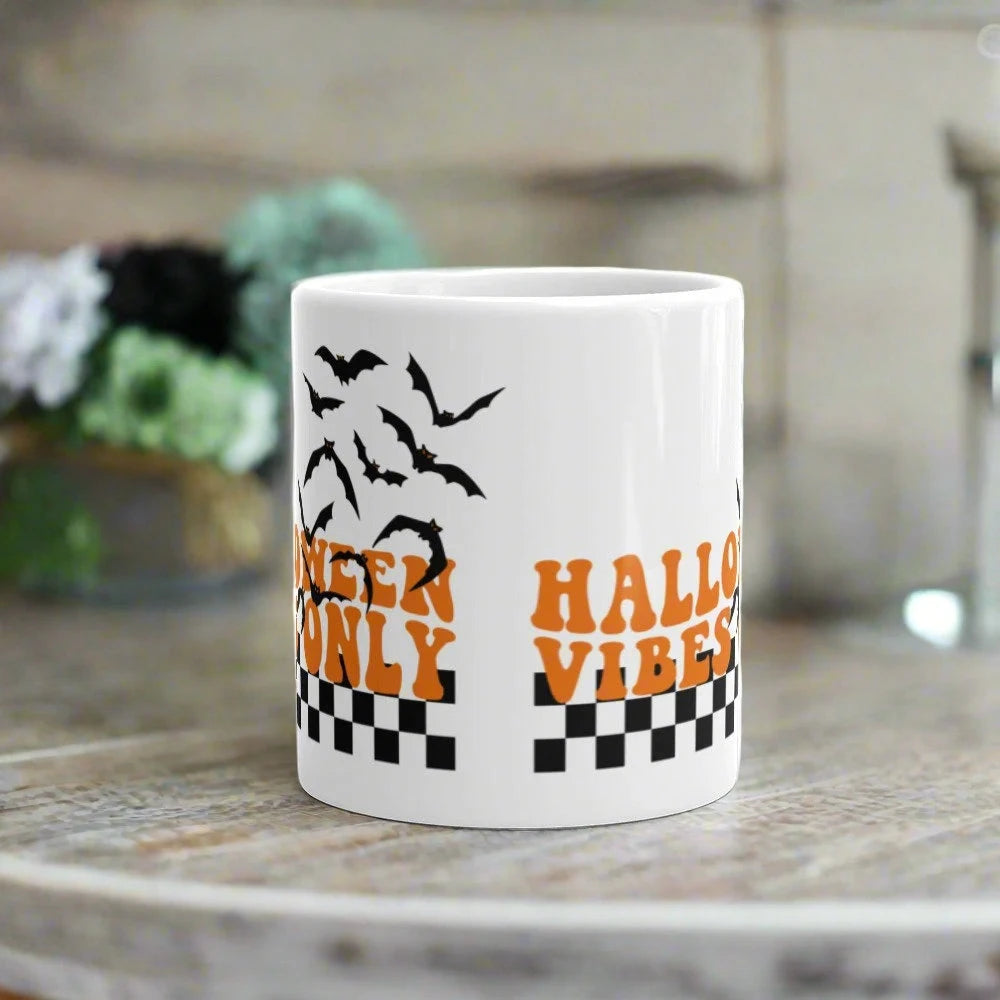 This ceramic glossy Halloween mug features a retro style print that says Halloween Vibes Only. There are flying bats and a black and white checkered print at the bottom.