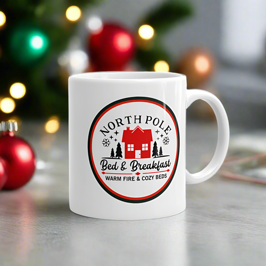 This ceramic glossy Christmas coffee mug says North Pole Bed & Breakfast Warm Fire & Cozy Beds. It features a little red house with trees and stars around it.