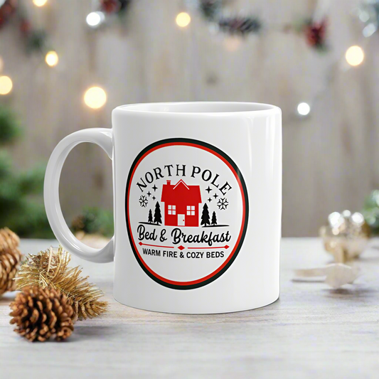 This ceramic glossy Christmas coffee mug says North Pole Bed & Breakfast Warm Fire & Cozy Beds. It features a little red house with trees and stars around it.