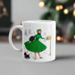 This ceramic glossy coffee mug features a midcentury retro Christmas illustration of a woman dressed in a green dress with a green hat, a black muff and red and white striped glove. She is walking her little black scottie and reaching into her mailbox. 