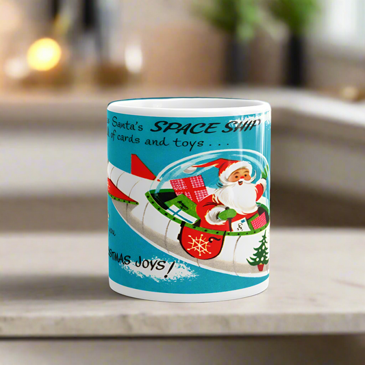 This ceramic glossy coffee mug featuring a  midcentury atomic retro Christmas illustration of Santa in a spaceship rocket. It says Here Comes Santa in a SPACE SHIP Full of cards and toys Zooming in to wish for you Lots of CHRISTMAS JOYS!