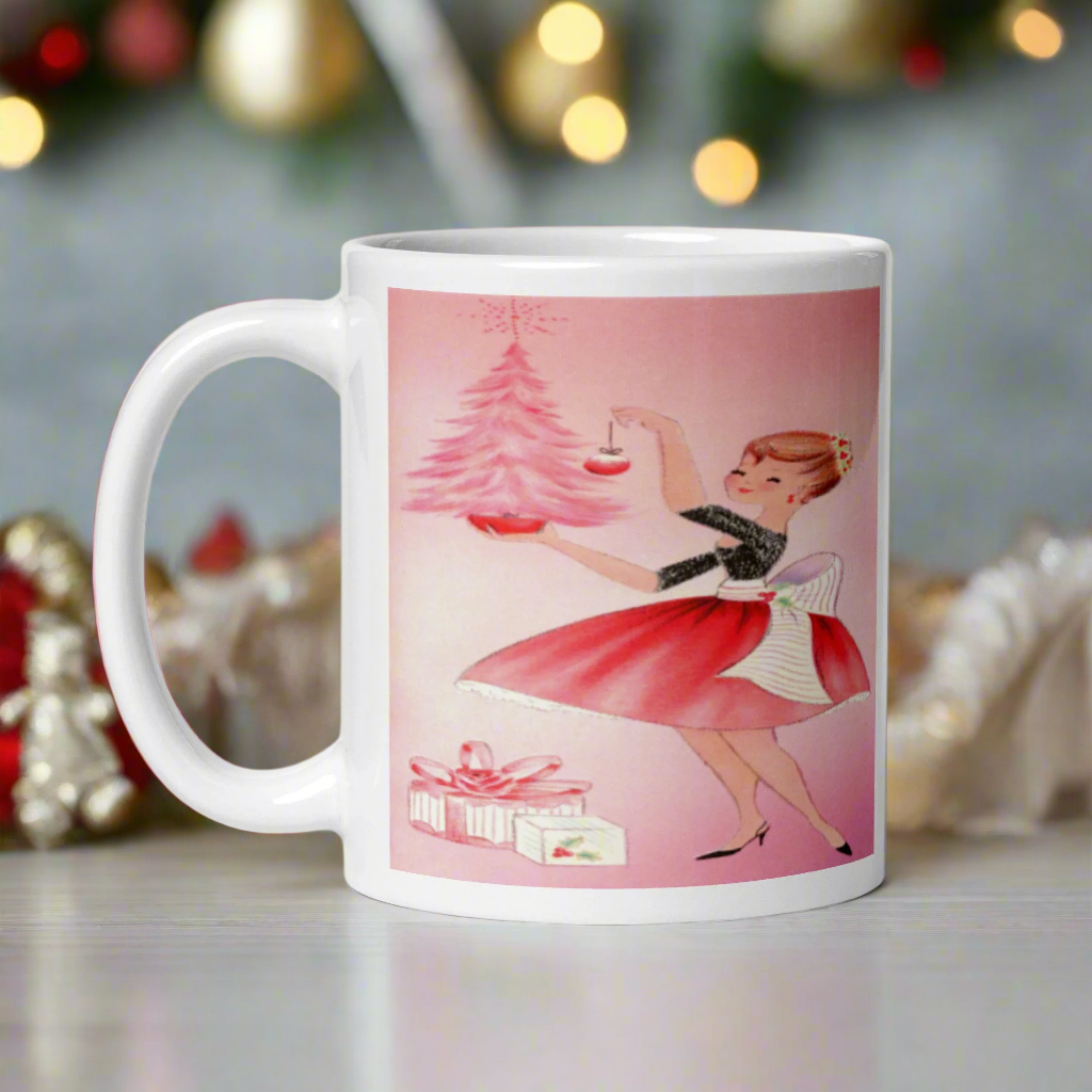 This glossy ceramic coffee mug features a midcentury retro Christmas illustration of a woman dressed in black and pink holding a small pink Christmas and ornament. There are a couple of presents and a pink background.