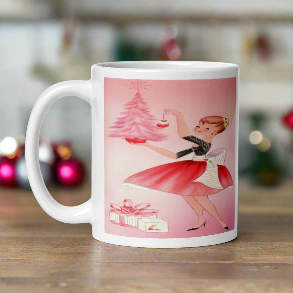 This glossy ceramic coffee mug features a midcentury retro Christmas illustration of a woman dressed in black and pink holding a small pink Christmas and ornament. There are a couple of presents and a pink background.