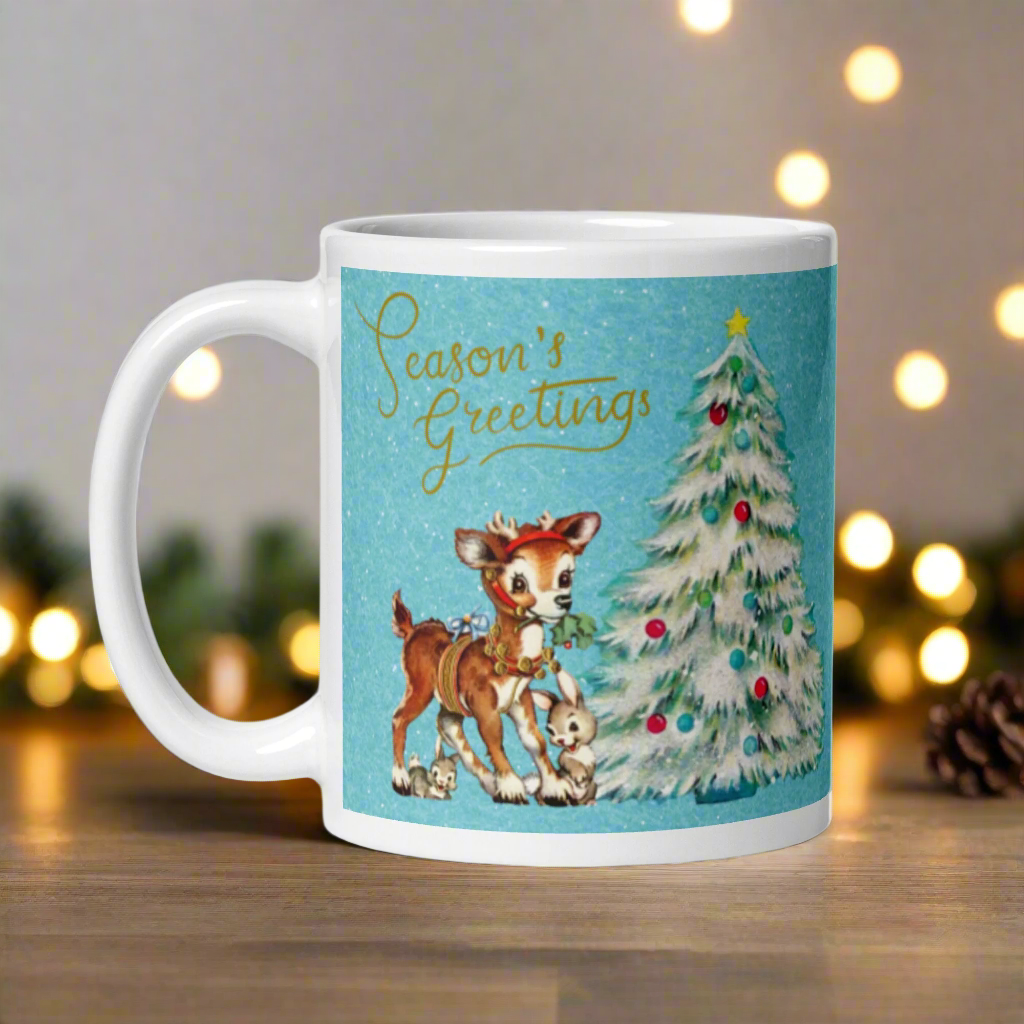 This glossy ceramic coffee mug features 
 Midcentury vintage artwork of an adorable reindeer with holly in her mouth, bunnies, and a Christmas Tree. There is gold lettering that says Season's Greetings and a blue felt background.