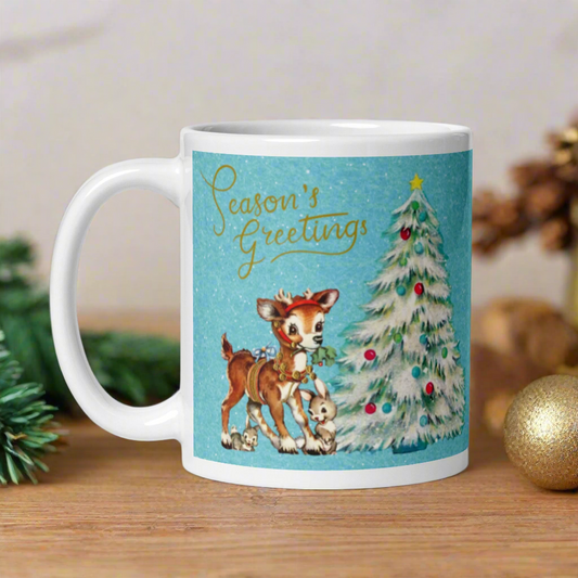 This glossy ceramic coffee mug features 
 Midcentury vintage artwork of an adorable reindeer with holly in her mouth, bunnies, and a Christmas Tree. There is gold lettering that says Season's Greetings and a blue felt background.