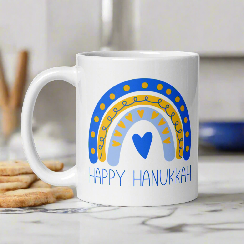 This ceramic glossy coffee cup says Happy Hanukkah and features a blue and yellow boho rainbow with a heart.