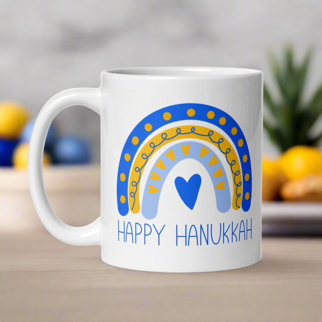 This ceramic glossy coffee cup says Happy Hanukkah and features a blue and yellow boho rainbow with a heart.