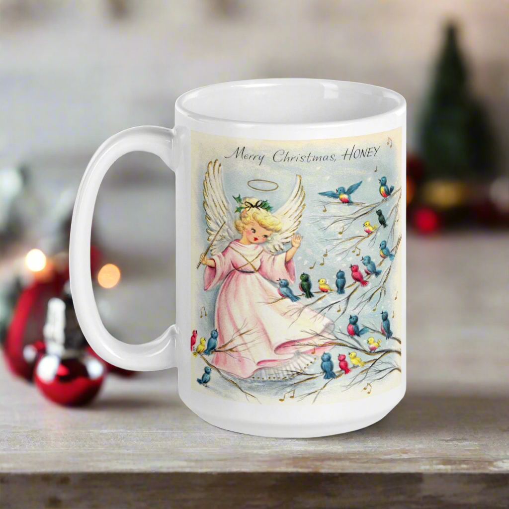 This ceramic glossy coffee mug features a retro Midcentury Christmas illustration of an angel dressed in pink directing blue, yellow and pink songbirds. At the top it says Merry Christmas HONEY