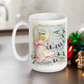 This ceramic glossy coffee mug features a retro Midcentury Christmas illustration of an angel dressed in pink directing blue, yellow and pink songbirds. At the top it says Merry Christmas HONEY