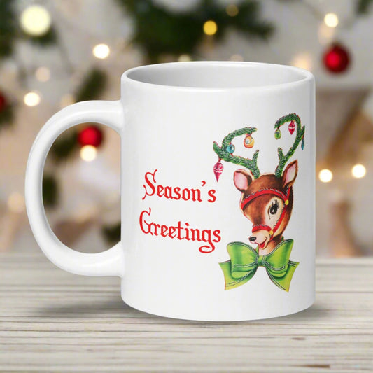 This ceramic glossy coffee mug features a midcentury vintage illustration of an adorable reindeer wearing a green bow with tinsel and ornaments hanging from her antlers. Red lettering says Season's Greetings.