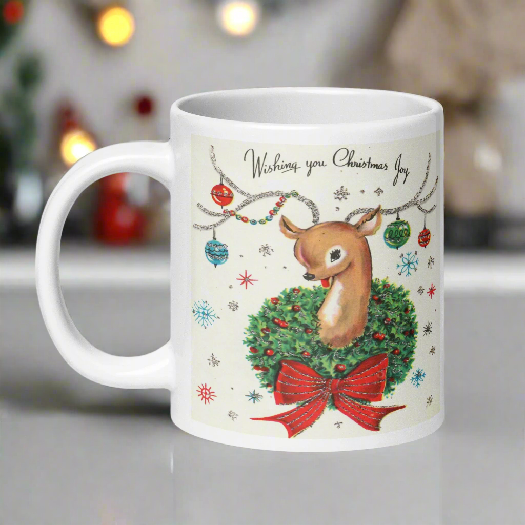 This ceramic glossy coffee mug is a midcentury christmas illustration that says Wishing you Christmas Joy. It features a reindeer with her tongue out wearing a Christmas wreath with a red bow. Her antlers are decorated with tinsel and  ornaments.