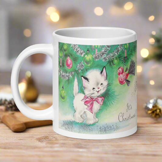 This ceramic glossy coffee mug features a midcentury vintage Christmas Illustration that says It's Christmas! There is an adorable white kitten wearing a pink bow underneath the christmas tree.