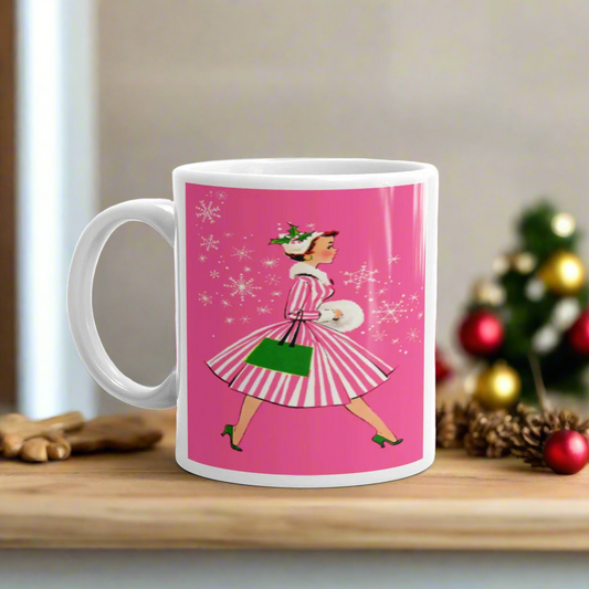 This ceramic glossy mug features a vintage Christmas illustration of a retro Midcentury housewife dressed in a pink and white striped dress with a green purse and heels. The background is pink and there are snowflakes. 